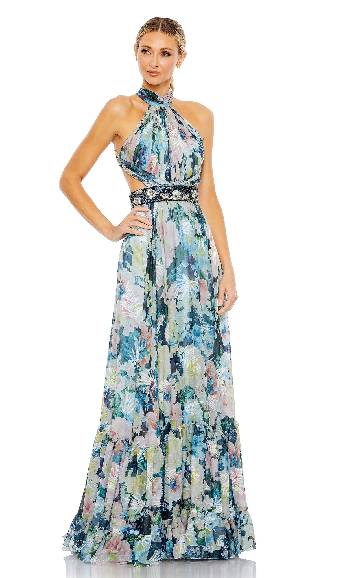 A woman models the Long Formal Dress 68089 by Mac Duggal, featuring pastel floral patterns, a ruched bodice, halter neckline, open back, and decorative waistband, standing against a plain white background.